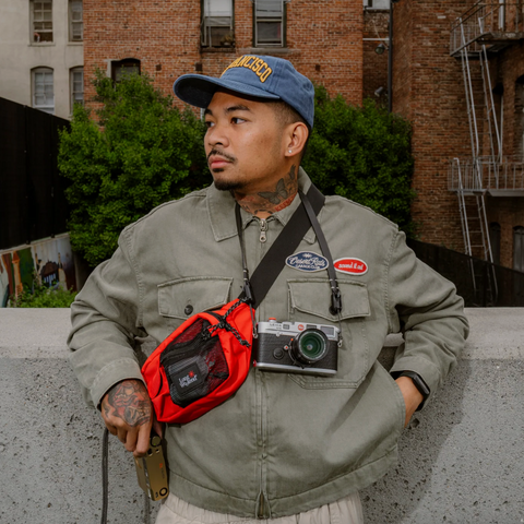 A man wearing a blue cap and an olive green jacket stands outdoors against a brick building background, wearing a red crossbody bag with a black pouch and a camera hanging from the strap.