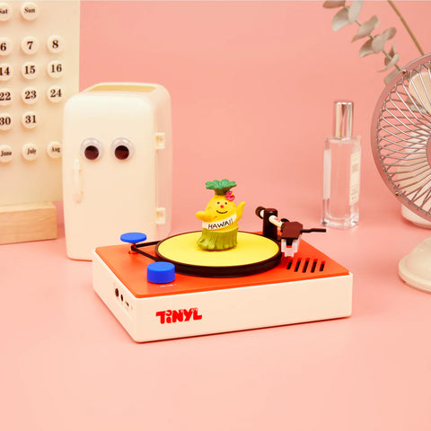 Toy record player with a small figure on a pink surface, featuring the brand 'TINYL'.
