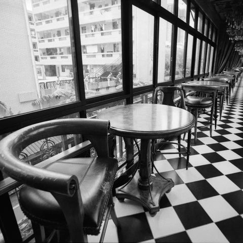 Black and white photograph of a long corridor with black-and-white checkered flooring, round tables, and leather chairs beside large windows overlooking a building exterior.