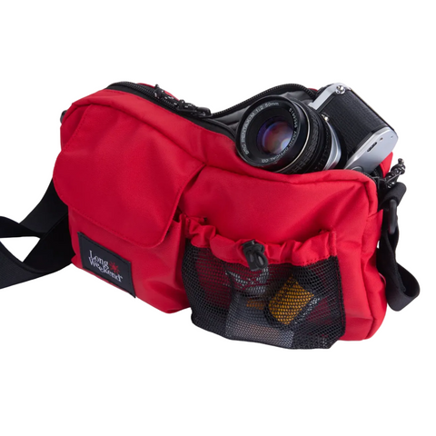 A red nylon camera bag with a black shoulder strap, shown holding a camera. The bag features a front mesh pocket containing a yellow item, a black brand patch on the lower left, and a silver camera partially tucked into the top compartment.