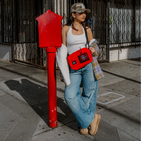 A woman stands on a city sidewalk next to a red mailbox, wearing a white tank top, blue jeans, beige boots, and a camouflage baseball cap, while carrying a bright red crossbody bag with a black star patch.
